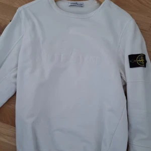 Vit sweatshirt från Stone Island - Säljer en stilren vit sweatshirt från Stone Island med klassisk logotyp-patch på ärmen. Tröjan har rund halsringning och långa ärmar. Perfekt för en clean och avslappnad look.