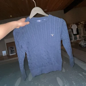 Mörkblå kabelstickad tröja från Gant - Mörkblå kabelstickad tröja från Gant med rund halsringning och diskret broderad logga på bröstet. Tröjan har långa ärmar och ribbade muddar vid ärmslut och nederkant. Perfekt för lager-på-lager och klassisk stil.