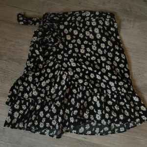 Svart blommig omlottkjol från ONLY - Supersöt svart omlottkjol från ONLY med vita och gula blommor. Kjolen har volangdetaljer och knyt i midjan. Perfekt för dig som gillar en feminin och lekfull stil. Använd endast en gång. 💗