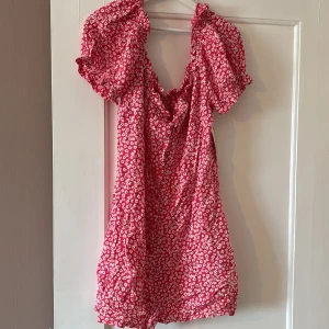 Röd blommig klänning från Nelly - Supersöt röd klänning från Nelly med vita blommor och korta puffärmar. Klänningen har en smockad överdel och volangdetaljer vid halsen. Perfekt för sommarens alla tillfällen!