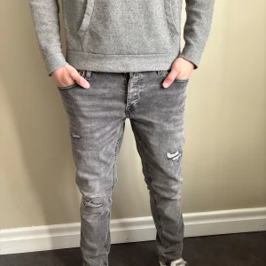 Grå slitna jeansbyxor från jack&jones - Säljer ett par grå jeansbyxor med slitna detaljer på benen. Byxorna har normal passform och klassisk femficksdesign. Perfekta för en avslappnad stil och passar till det mesta. Snygga sömmar på bakfickorna och dragkedja i gylfen.