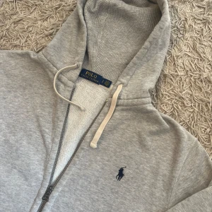 Grå hoodie med dragkedja från Polo Ralph Lauren grey grisch sweater  - Snygg ljusgrå hoodie från Polo Ralph Lauren med dragkedja och huva. Tröjan har en broderad marinblå logga på bröstet och vita snören i huvan. Perfekt för en avslappnad och stilren look. Storlek S men mått kan såklart skickas vid osäkerhet. Använd men inga defekter. Postas antigen samma dag som köp eller dagen därpå🙌💯