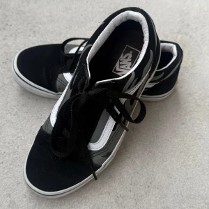 Svarta och vita Vans sneakers - Klassiska Vans sneakers i svart och vitt med vit sula och svarta skosnören. Skorna har den ikoniska sidoremsan i vitt och är låga i modellen. Perfekta för en avslappnad och stilren look.