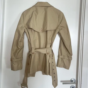 Beige trenchcoat från InWear - Snygg beige trenchcoat från InWear med klassisk krage, dubbelknäppning och bälte i midjan 🩷 Jackan har långa ärmar med spänne vid ärmslut och flera fickor framtill. Perfekt för en stilren ock klassisk look!