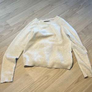 Beige stickad tröja från Vero Moda - Säljer en enkel och stilren beige stickad tröja från Vero Moda. Tröjan har rund halsringning, långa ärmar och ribbade muddar vid ärmslut och nederkant. Perfekt för lager-på-lager under kyligare dagar.