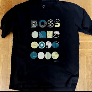 Svart t-shirt från BOSS med grafiskt tryck - Svart t-shirt från BOSS med unikt grafiskt tryck i flera färger på framsidan. Trycket består av olika varianter av BOSS-loggan i blått, beige, vitt och gult. Klassisk passform och rund halsringning.