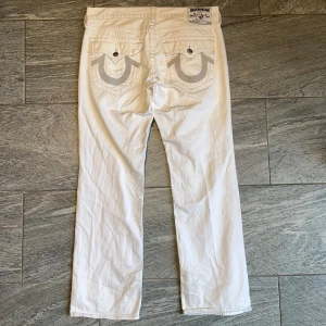 Vita jeansbyxor från True Religion - Säljer ett par vita jeansbyxor från True Religion med raka ben och klassiska bakfickor med broderad logga. Byxorna har knappar och dragkedja framtill samt detaljerade sömmar. Perfekta för dig som vill sticka ut med en clean look.