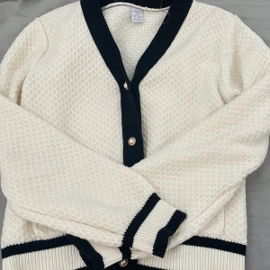 Vit kofta med svarta detaljer - Säljer en vit stickad kofta med svarta ribbade kanter och knappar framtill. Koftan har lång ärm och ett klassiskt, elegant utseende med strukturstickat mönster. Perfekt att slänga över en topp för en snygg look.