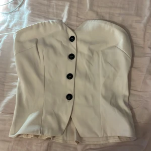 Vit tubtopp med knappar - Säljer en stilren vit tubtopp med fyra svarta knappar framtill. Toppen har en figurnära passform och en asymmetrisk skärning upptill. Perfekt att styla till olika outfits!