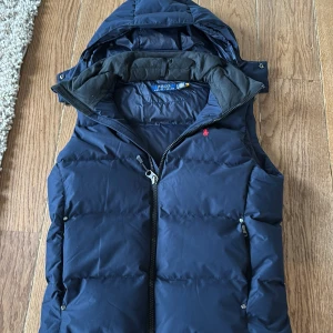 Mörkblå dunväst från Polo Ralph Lauren - Snygg mörkblå dunväst från Polo Ralph Lauren med avtagbar huva och dragkedja framtill. Den är bara andvänd 4 gånger. Ny pris är 4500