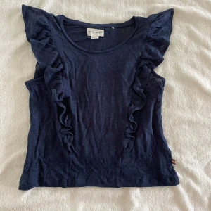 Mörkblå ärmlös topp med volang från Holly & Whyte - Supersöt mörkblå topp från Holly & Whyte med volangdetaljer längs axlarna. Ärmlös modell i mjukt material, perfekt till sommaren. Enkel rund halsringning och liten tygdetalj i sidan. Den är ganska genomskinlig - Jag har 20% bundle rabatt om du köper 2 plagg 🦋