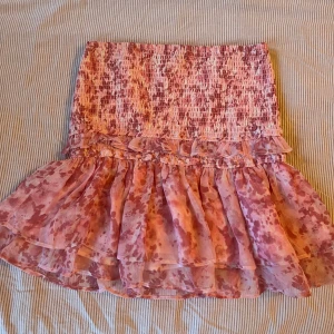 Rosa volangkjol från Gina Tricot - Supersöt rosa kortkjol från Gina Tricot med smock upptill och volangdetalj. Kjolen har ett rosa mönster och är gjord i ett lätt, luftigt material som passar perfekt till varmare dagar.