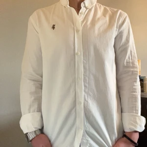 Vit skjorta från U.S. Polo Assn. - Klassisk vit skjorta från U.S. Polo Assn. med knappar framtill och broderad logga på bröstet. Skjortan har lång ärm och en stilren, tidlös design som passar till många tillfällen. Tillverkad i bomull för en bekväm känsla.