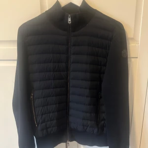 Moncler cardigan hybrid  - Säljer denna extremt snyggra och stilrena Moncler cardigan hybrid i storlek M (passar S). Skicket är 9/10. Det finns två små hål på båda ärmarna nere där händerna sticker ut. Dragkedjan och sömmarna i ärmarna är nyfixade  hos moncler för 1 år sedan. Nypris är runt 13,000kr. Mitt pris 2995kr. Utmärkt pris och snygg jacka som passar till alla outfits😁