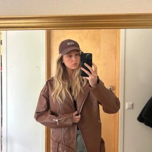 Brun skinnjacka  - Säljer en brun skinnjacka med coola dragkedjedetaljer och bälte i midjan. Jackan har en oversized passform och långa ärmar, perfekt att styla med jeans och keps för en avslappnad look.