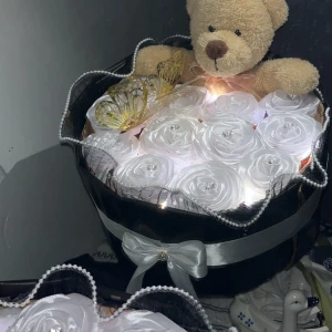 Bukett med vita rosor, nalle och fjäril - En söt bukett med konstgjorda vita rosor, dekorerad med en mjuk nallebjörn och en guldfärgad fjäril. Buketten är inslagen i svart och silverfärgat papper med en stor vit rosett framtill. Perfekt som present eller dekoration.