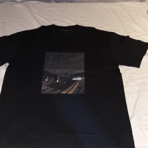 Svart t-shirt med biltryck från Natone  - Svart t-shirt från Natone med ett stort tryck på framsidan som föreställer en klassisk bil som kör på en väg under en stjärnklar natthimmel. T-shirten har rund hals och korta ärmar. Perfekt för dig som gillar retrostil och coola motiv. OANVÄND