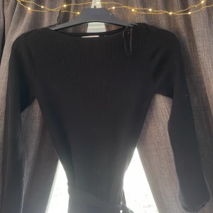 Svart ribbad klänning med knytband från Holly & Whyte - Säljer en svart ribbad långärmad klänning från Holly & Whyte med knytband i midjan. Klänningen har en rund halsringning och markerad midja för en snygg siluett. Perfekt för dig som gillar stilrena och bekväma plagg.