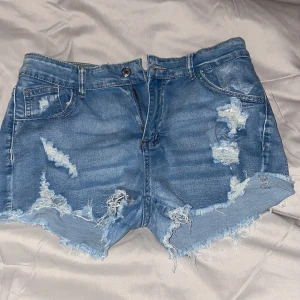 Blå jeansshorts med slitningar och fransar - Snygga blå jeansshorts med slitna detaljer och fransig kant nertill. Klassisk femficksmodell med knapp och dragkedja framtill. Perfekta för sommaren och ger en avslappnad look.