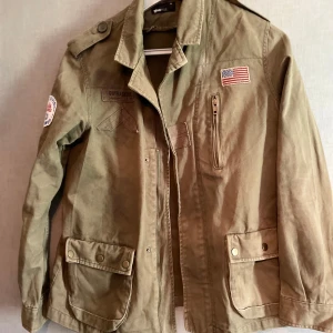 Olivgrön militärinspirerad jacka från Gina Tricot - Säljer en olivgrön militärjacka från Gina Tricot med patchar, bland annat amerikansk flagga och Air Guards-märke på ärmen. Jackan har flera fickor med knappar och dragkedja framtill. Perfekt för dig som gillar utility-stil.