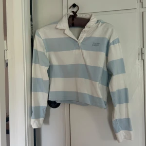 Randig rugbytröja - Säljer en rugbytröja från Levi's i vita och ljusblåta breda ränder. Tröjan har krage, knäppning vid halsen och broderad logga på bröstet. Perfekt för en avslappnad och sportig stil. Använd typ en gång🥰så fin nu till våren och sommaren!