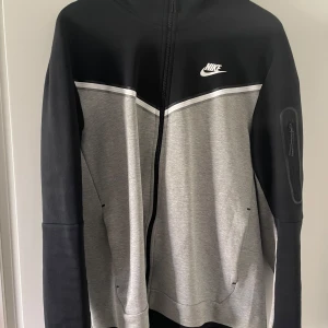 Svart och grå hoodie från Nike - Säljer en snygg hoodie från Nike i svart och grått med dragkedja framtill och vit logga på bröstet. Tröjan har huva, långa ärmar och praktiska fickor. Perfekt för en sportig och avslappnad stil. Samt har byxorna till hoodien och tsm blir det 1000kr byxor L  pris kan sänkas 