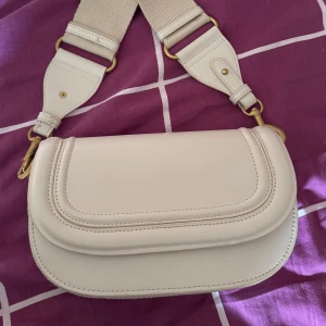 Vit axelväska från Zara - Säljer en stilren vit axelväska från Zara med bred, justerbar axelrem och guldfärgade metalldetaljer. Väskan har ett huvudfack med innerficka med dragkedja. Perfekt för att bära det viktigaste med stil.