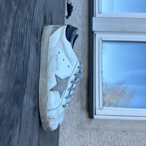 Vita sneakers från Golden Goose med grå stjärna - Säljer ett par vita sneakers från Golden Goose med grå stjärndetalj på sidan och svart hälparti. Skorna har snörning och en platt sula. Perfekta för dig som gillar en stilren och trendig look. Skadade där fram som man ser på bild 3.  Pris kan absolut diskuteras.  De är limmat så de håller helt okej men går nog att fixa. Vet inte om de e fejk eller inte. 