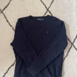 Mörkblå stickad tröja från Polo Ralph Lauren - Säljer en klassisk mörkblå stickad tröja från Polo Ralph Lauren. Tröjan har kabelstickat mönster, rund halsringning och långa ärmar. Perfekt för lager-på-lager och stilren look.