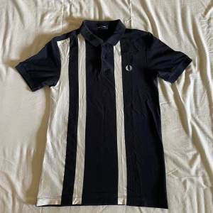 Mörkblå, svart och vit randig pikétröja från Fred Perry - Snygg marinblå pikétröja från Fred Perry med vita vertikala ränder framtill och klassisk krage. Tröjan har korta ärmar och knäppning vid halsen. Bara använd ett fåtal gånger. 