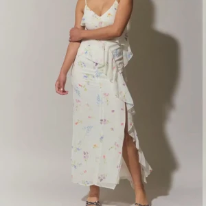 Vit blommig långklänning från Gina Tricot - Helt ny Superfin vit långklänning från Gina Tricot med tunna axelband och blommigt mönster i pastellfärger. Klänningen har volangdetaljer och en slits framtill, perfekt för sommarens alla tillfällen.