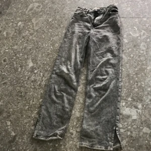 Grå tvättade jeansbyxor från Lindex - Säljer ett par grå tvättade jeansbyxor från Lindex med raka ben och slitsar nertill. Byxorna har klassisk femficksdesign och normal passform. Perfekta för en avslappnad stil.
