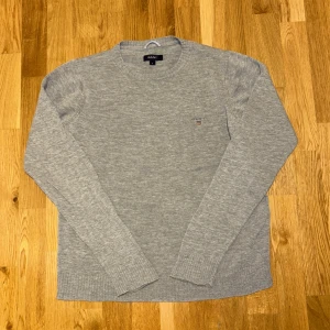 Grå stickad tröja från GANT - Säljer en stilren grå stickad tröja från GANT med rund halsringning och diskret logga broderad på bröstet. Tröjan har långa ärmar och ribbade muddar vid ärmslut och nederkant. Perfekt för lager-på-lager under kyliga dagar.