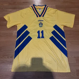 Svensk landslagströja Adidas Brolin 11 - Gul kortärmad fotbollströja från Adidas med blå detaljer och nummer 11 på bröstet och ryggen. Svenska landslagets emblem på framsidan och namnet Brolin på ryggen. Klassisk v-ringad krage med blå och gula ränder.