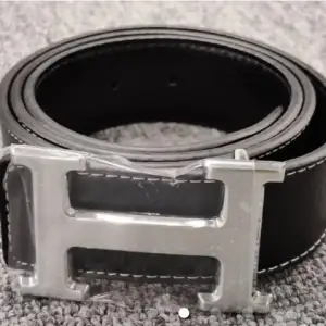Snyggt svart bälte med ett stort silverfärgat H-format spänne. Bältet har en stilren design med vita sömmar längs kanterna. Perfekt för att ge en elegant touch till din outfit.