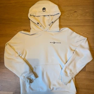 Vit hoodie från Sail Racing - Säljer en stilren vit hoodie från Sail Racing med logga på bröstet och huva med dragsko. Tröjan har en stor magficka och är tillverkad i en mjuk bomulls- och polyestermix. Perfekt för en avslappnad look.
