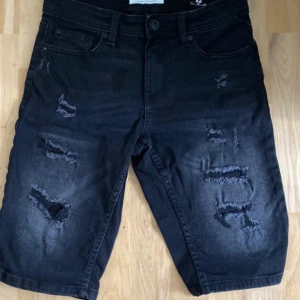 Svarta jeansshorts med slitningar från FSBN - Svarta jeansshorts från FSBN med flera slitna detaljer framtill. Klassisk femficksmodell med knapp och dragkedja i gylfen. Perfekta för en avslappnad stil.