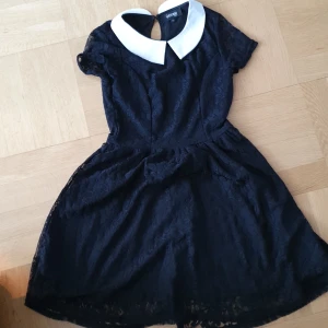 Svart spetsklänning med vit krage från Gothicana - Säljer en svart klänning från Gothicana med vit, stor krage och korta ärmar. Klänningen har spetsmönster och går ungefär till knäna på mog som är 160. Är knappt använd.