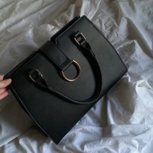 Svart handväska med gulddetaljer - Elegant svart handväska med stilrena gulddetaljer. Väskan har en praktisk design med två handtag och en dekorativ ring framtill. Perfekt för att bära dina dagliga nödvändigheter med stil.