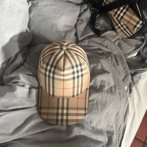 Beige rutig keps från Burberry - Snygg keps från Burberry i klassiskt beige rutigt mönster med svarta, röda och vita linjer. Justerbar rem baktill och broderad Burberry-logga. Perfekt accessoar för en trendig look.