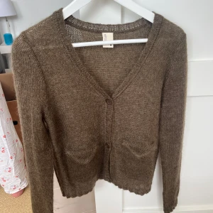 Brun stickad kofta från Rützou - Mysig brun stickad kofta från Rützou med v-ringning och två knappar framtill. Koftan har långa ärmar och två små fickor framtill. Perfekt för lager-på-lager och enkel att matcha med olika outfits.