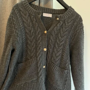 Mörkgrå stickad kofta från Holly & Whyte - Mysig mörkgrå stickad kofta från Holly & Whyte med grovt flätmönster framtill, två stora fickor och guldfärgade knappar. Perfekt för lager-på-lager och har ribbade muddar vid ärmslut och nederkant.