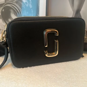 Svart axelväska från Marc Jacobs - Snygg svart axelväska från Marc Jacobs med guldfärgad logga framtill. Väskan har två dragkedjefack, justerbar axelrem och ett praktiskt ytterfack på baksidan. Perfekt för att bära det viktigaste med stil. Knappt använd