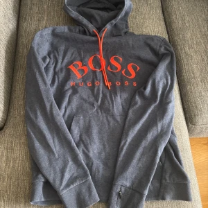 Blå hoodie från Hugo Boss med orange tryck - Snygg grå hoodie från Hugo Boss med stort orange BOSS-tryck. Mjuk bomullskänsla och klassisk passform med långa ärmar och huva. Perfekt för en avslappnad och stilren look. St L men passar M bättre.
