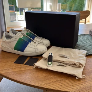 Valentino sneakers med blå och grön detalj - Säljer ett par vita Valentino sneakers med en bred blå och grön rand över mitten. Skorna har rund tå, platt sula och klassisk snörning. Märkeslogga på plösen och kommer med originalkartong, dustbags och extra skosnören.