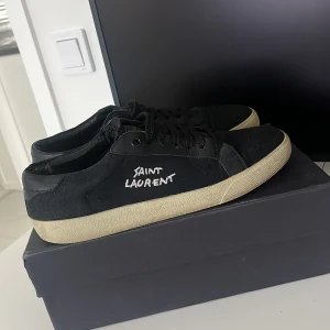 Saint Laurent skor - Svarta låga sneakers från Saint Laurent med vit logotyp på sidan och vit sula. Skorna har snörning och en klassisk rund tå. Perfekta för dig som gillar stilrena och exklusiva sneakers med streetkänsla. Man får med dustbag och box. Det finns en liten defekt uppe vid snörena på vänster skon.