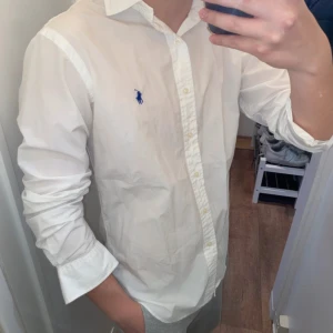 Polo Ralph Lauren (Vit skjorta) - Säljer en klassisk vit skjorta från Polo Ralph Lauren med den ikoniska broderade loggan i blått på bröstet. Skjortan har länga ärmar och knappar framtill. Perfekt för en stilren look. Den är i underbar skick och redo för en ny ägare! Skjortan är size M!