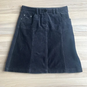 Svart manchesterkjol från Cometex - Säljer en svart manchesterkjol från Cometex i storlek 38. Kjolen har klassisk femficksdesign, bälteshällor och knappstängning framtill. Perfekt för dig som gillar en tidlös och enkel stil. Går även att klippa av och sy om till en kortare kjol.