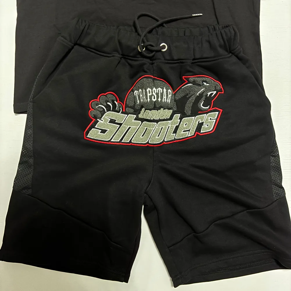 Snygg svart t-shirt och shorts från Trapstar med ett coolt 'London Shooters'-tryck i grått och rött. Seten har korta ärmar och en normal passform. Perfekt för en avslappnad stil. Båda ( T-shirt och Shorts följer med ). T-paidat.