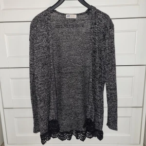 Gråmelerad kofta med spetskant från H&M - Säljer en gråmelerad kofta från H&M med lång ärm och snygg svart spetsdetalj längst ner. Perfekt att slänga över en topp för en chill och stilren look. Koftan är öppen framtill och har en mjuk, stickad känsla.